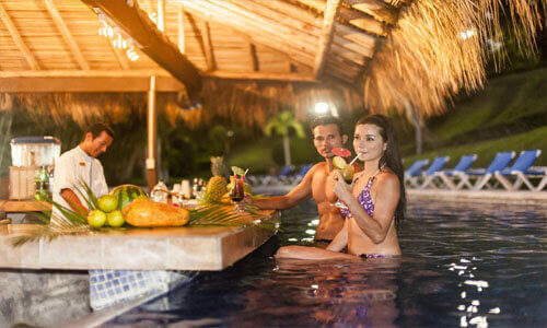 Picture of a hotel’s outdoor pool bar serving drinks in Costa Rica.  The picture shows two guests under a grass hut having drinks.