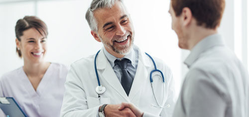 Picture of a doctor shaking hands with a male patient.  The picture shows a doctor with a white coat and stethoscope around his neck, smiling with a nurse standing behind him.