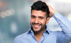 Picture of a man, happy with his hair transplant procedure he had at Top Plastic Surgeons in beautiful San Jose, Costa Rica.  The man is wearing a blue shirt with an open collar and holding his left hand to his head.