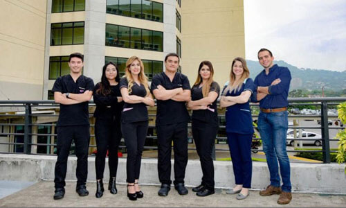Picture of a smiling medical team at CIMA Hospital  in San José, Costa Rica.  The picture shows seven members of the medical group standing with arms folded with CIMA Hospital behind them.