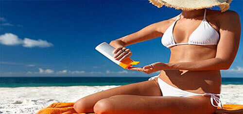 Picture of an attractive woman on a beautiful Costa Rica beach, applying suntan lotion.  The woman has on a two-piece white bathing suit and she is sitting on a beach towel with a beautiful blue Costa Rica sky in the background.